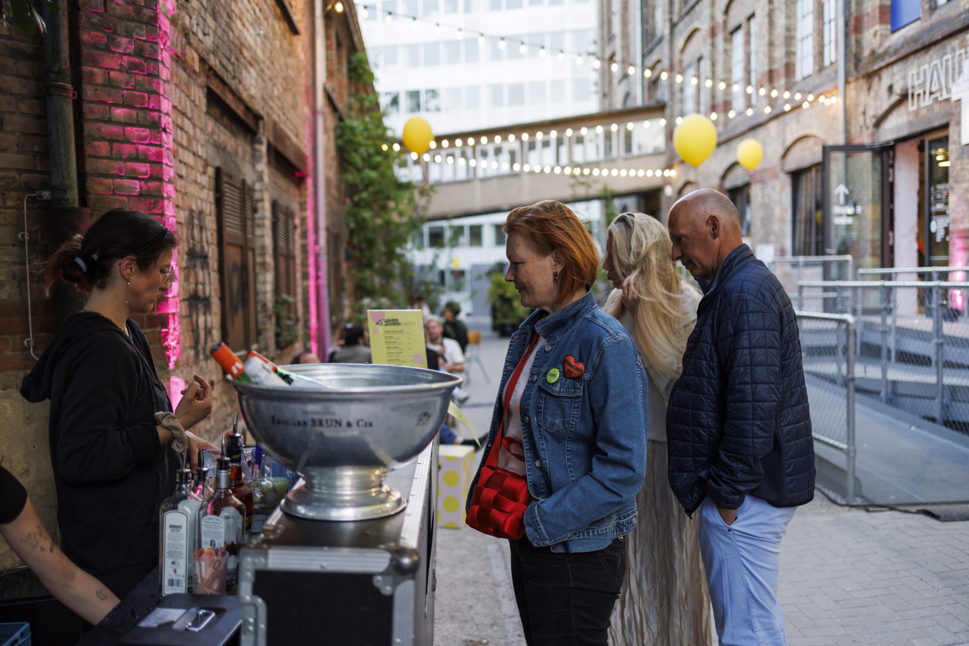 Eine Frau am Bartresen unterhält sich mit einer anderen Frau; im Hintergrund sind weitere Personen in festlicher Atmosphäre.