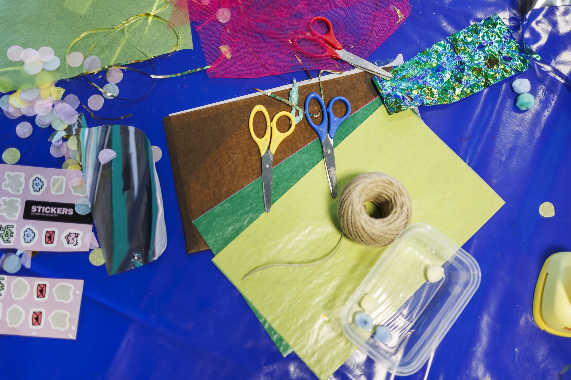 Bastelmaterialien wie buntes Papier, Scheren, Kleber und Dekoteile auf einem blauen Tisch.