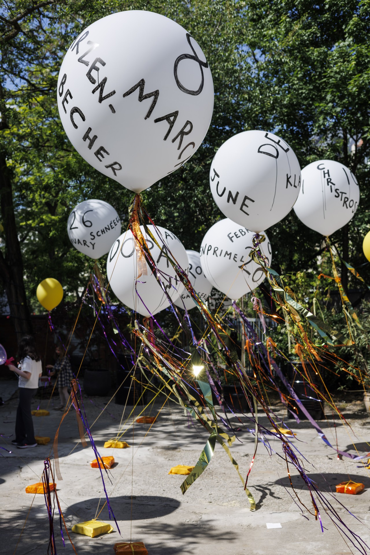 Bunte Luftballons mit Namen und Daten, geschmückt mit bunten Bändern, in einem sonnigen Park.