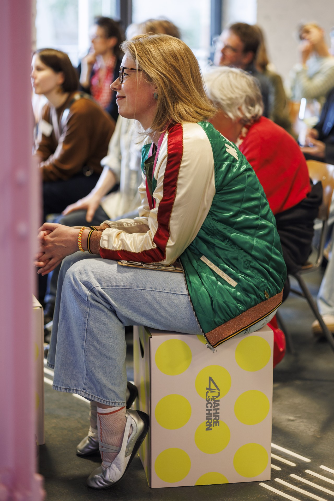 Frau mit blonden Haaren sitzt auf einem bunten Hocker, trägt eine grüne Jacke und schaut aufmerksam.