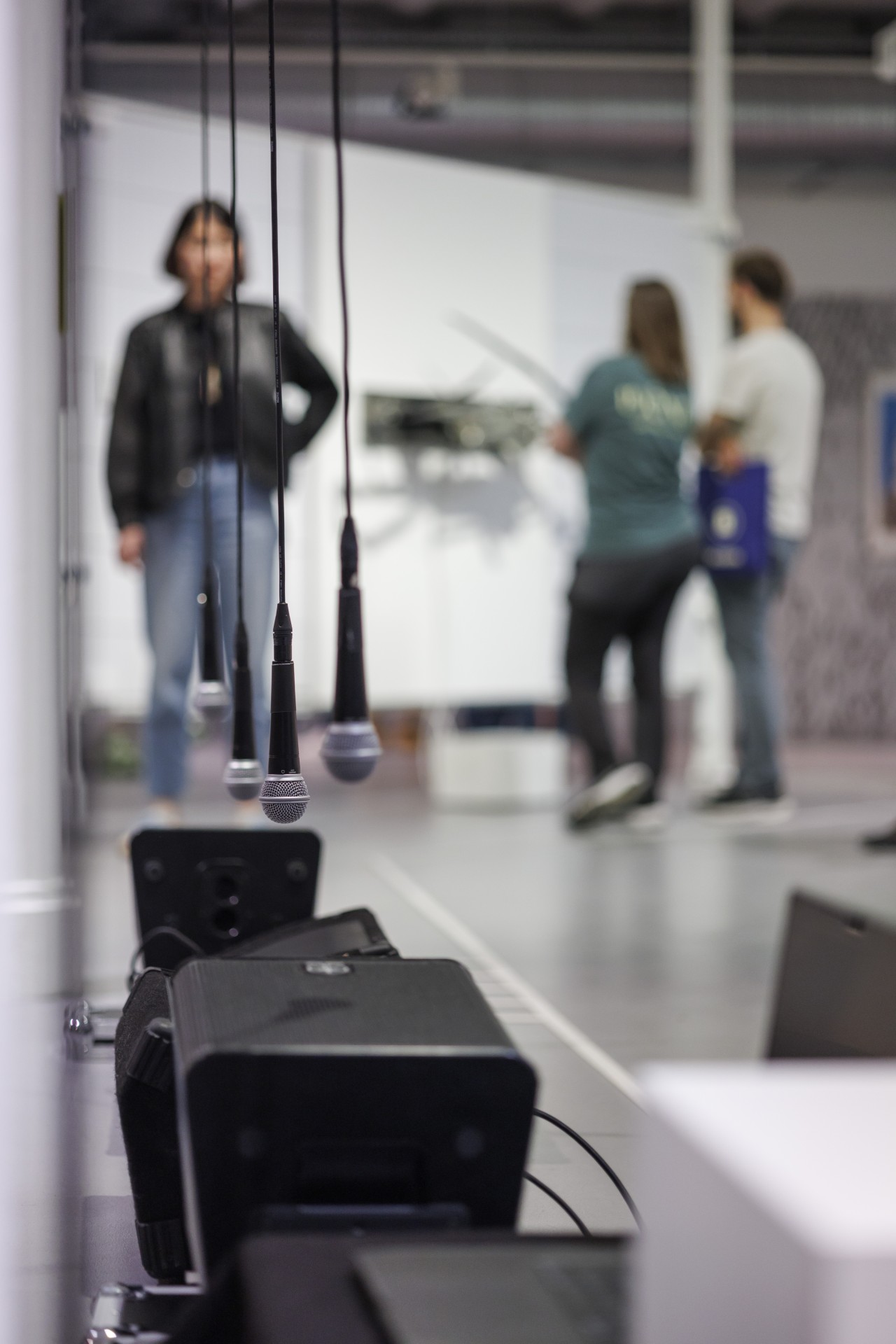 Mikrofone hängen in einem Ausstellungsraum, während Besucher im Hintergrund miteinander interagieren.
