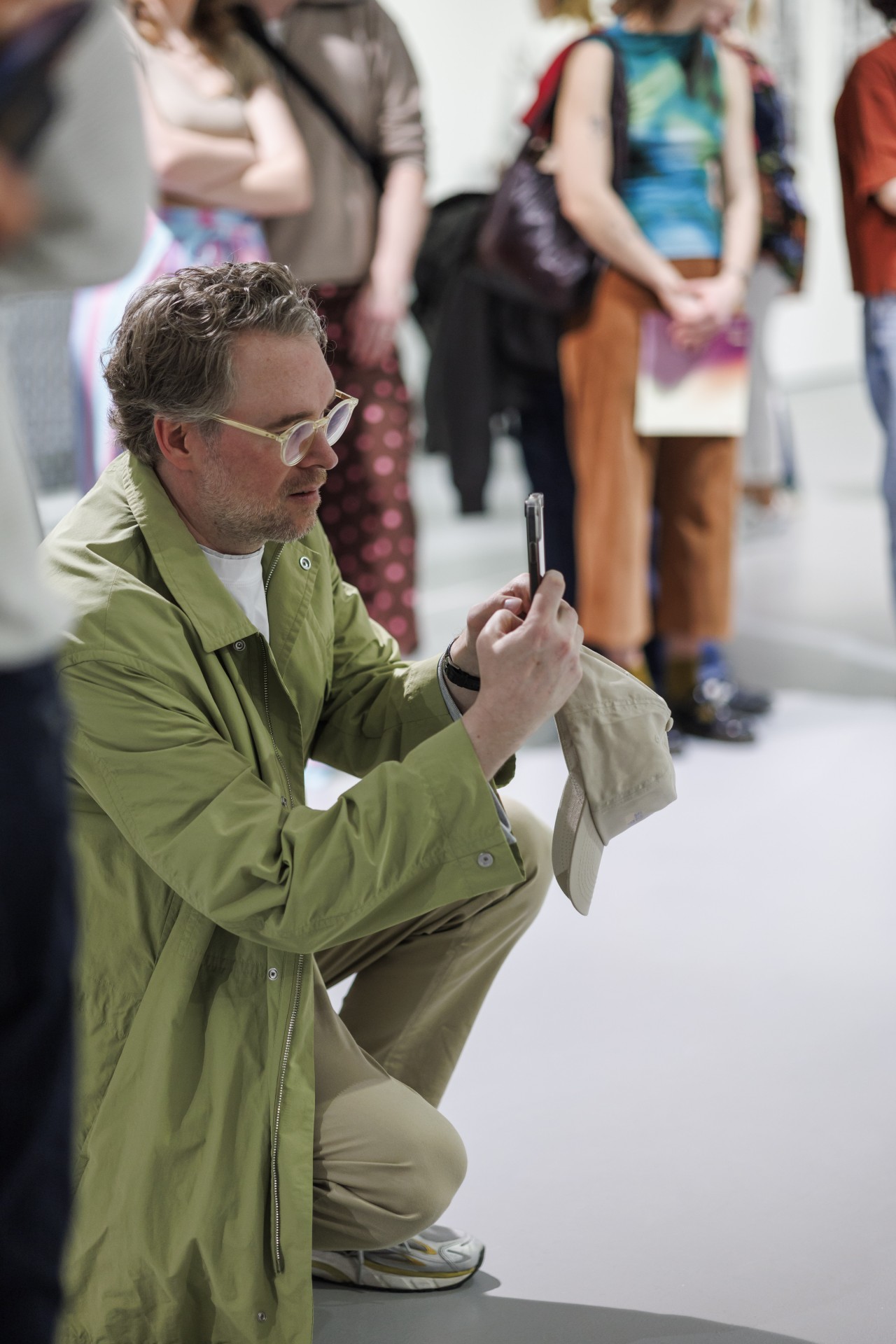 Mann im grünen Mantel kniet und fotografiert mit seinem Handy, umgeben von Besuchern in einer Galerie.