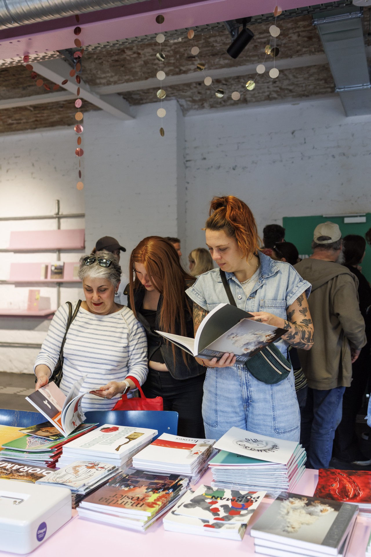 Drei Frauen blättern in Büchern an einem Tisch mit Zeitschriften in einem hellen, modernen Raum.