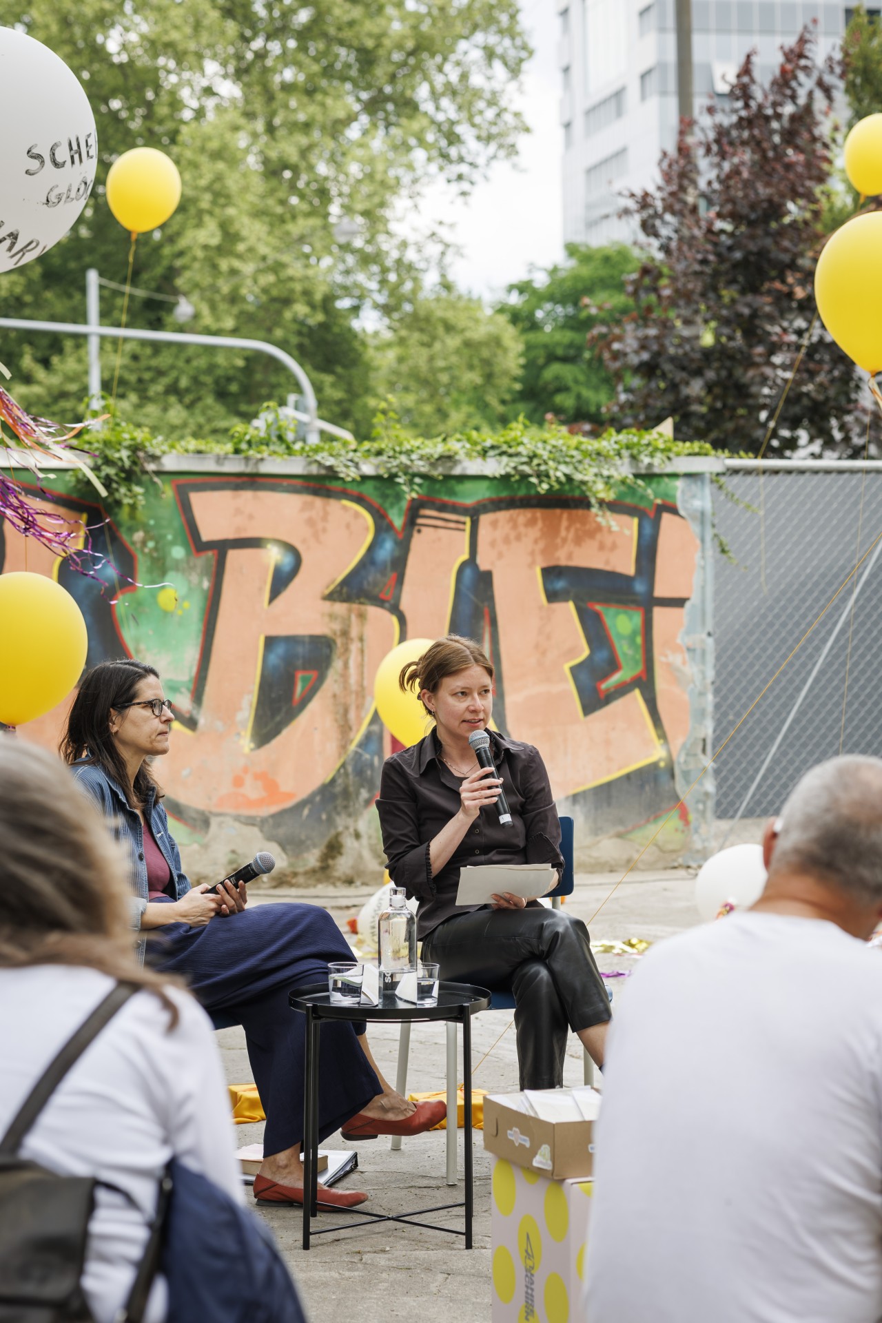 Zwei Frauen unterhalten sich, umgeben von gelben Ballons und einer bunten Graffiti-Wand.