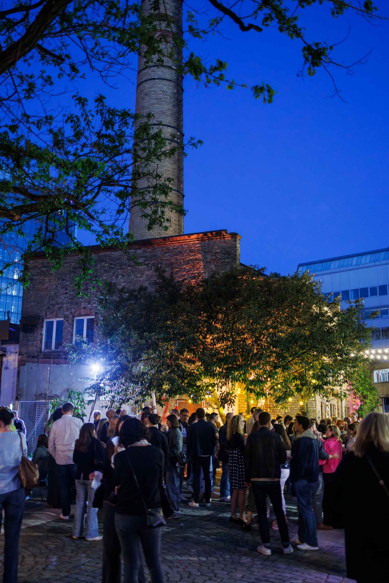 Lively evening atmosphere in front of an old factory, surrounded by trees and evening lighting against a blue sky.