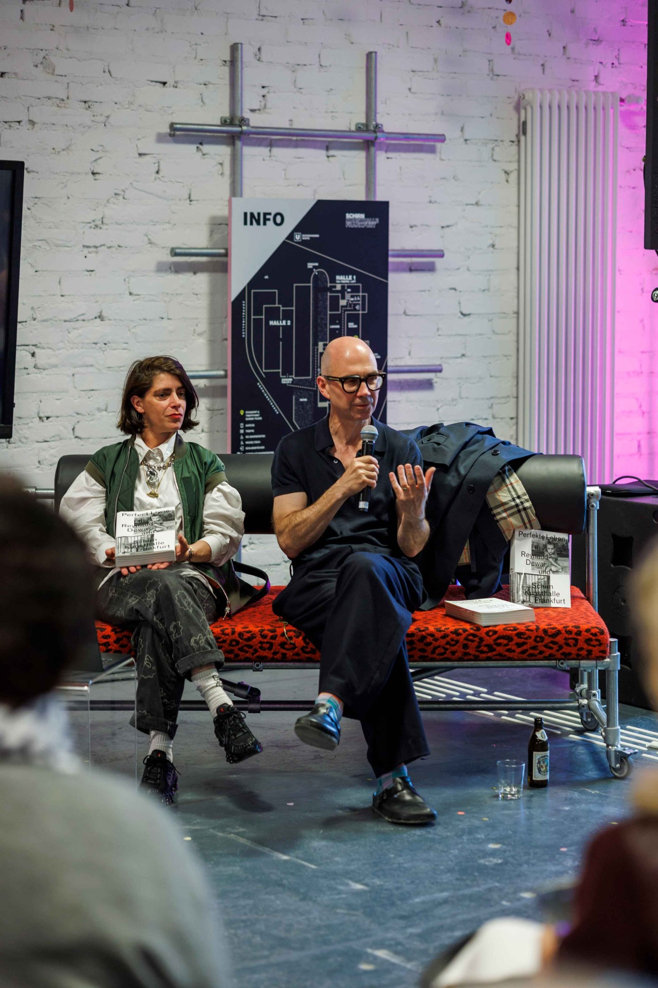 Two people are discussing on a sofa, surrounded by books and a poster in the background.
