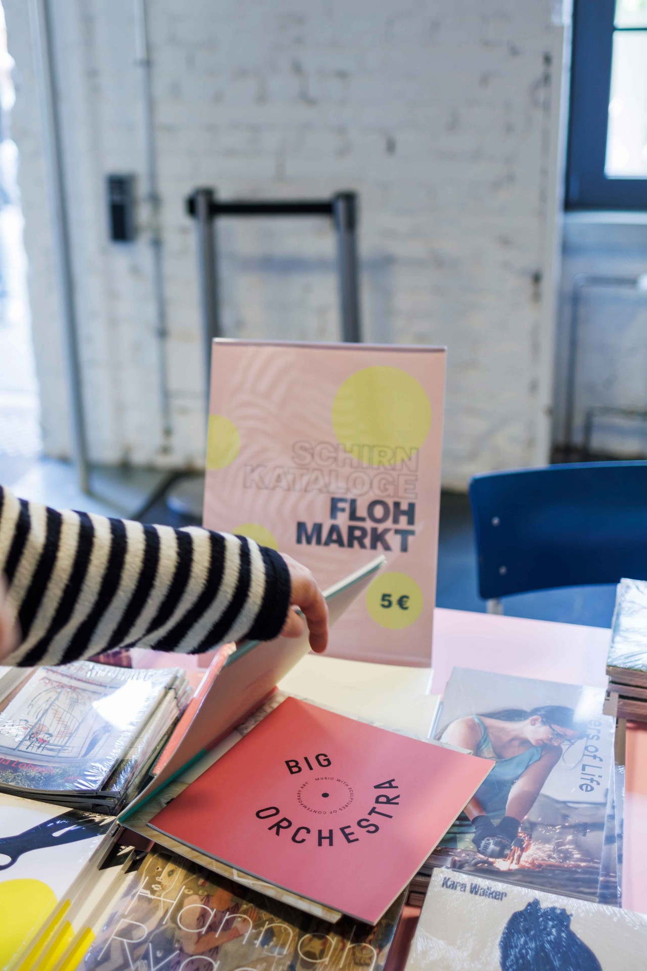 A stand with books and a pink flea market sign. A hand flips through catalogs. Colorful, creative atmosphere.