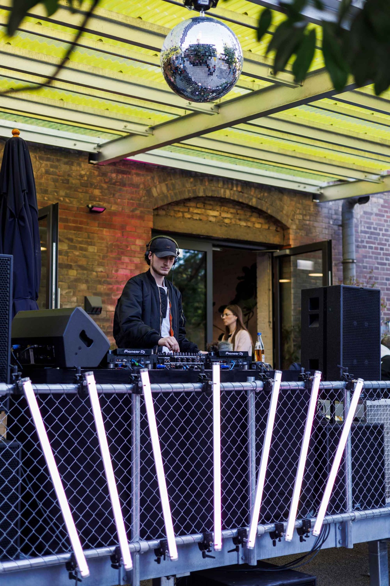 DJ at a mixing console under a large disco ball, surrounded by lights and a relaxed atmosphere.