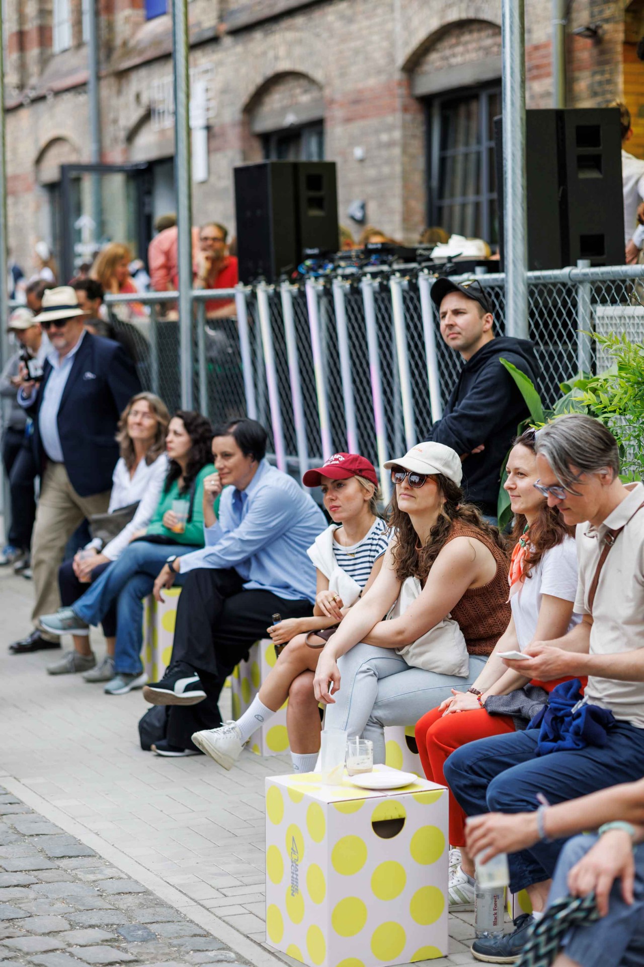 A group of people sits relaxed on colorful boxes in an urban setting during an event.