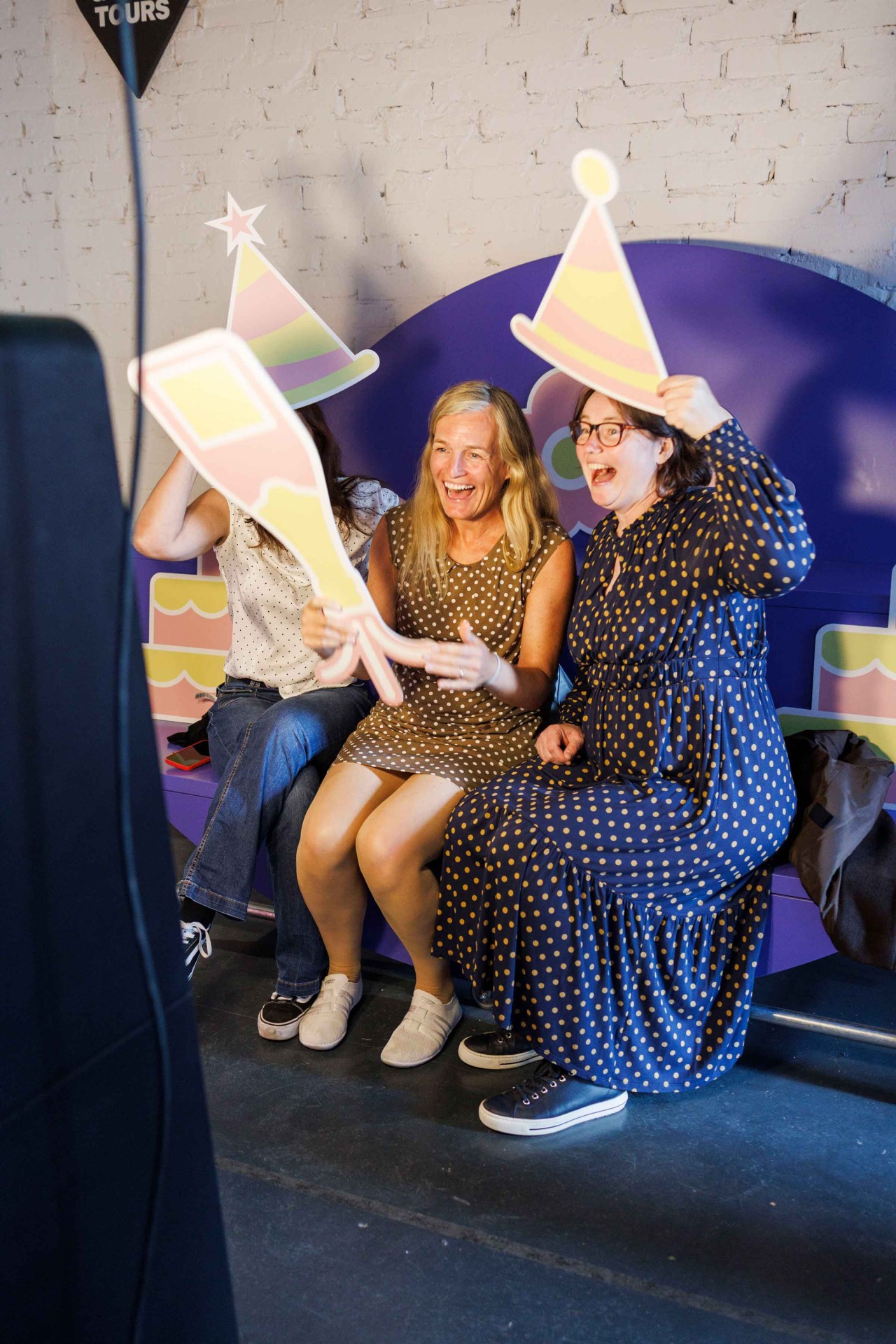 Three women in a festive atmosphere with party hats, laughing and posing in front of a colorful wall.
