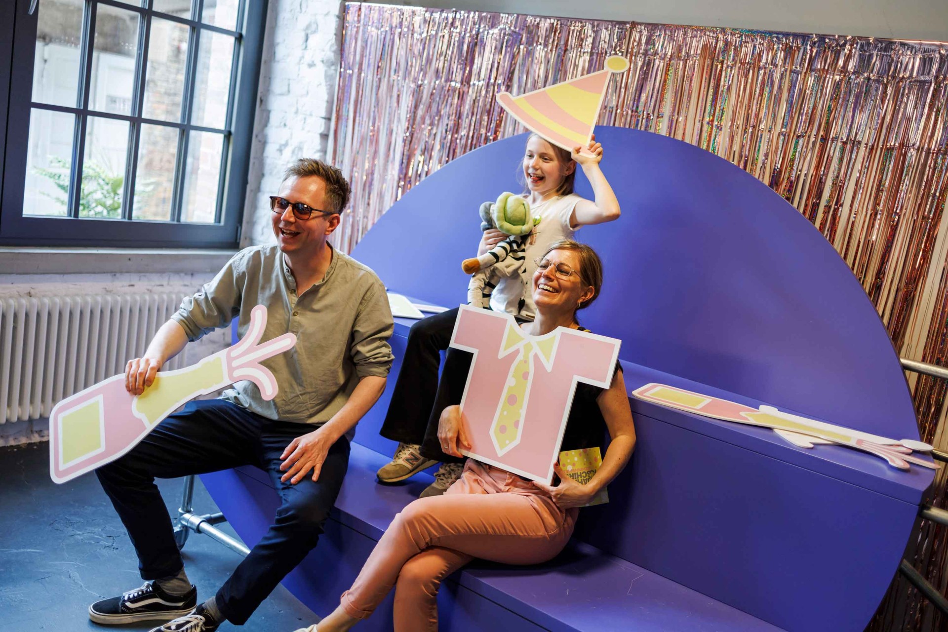 Three people happily pose with colorful props in a festive room. A child holds a stuffed animal.