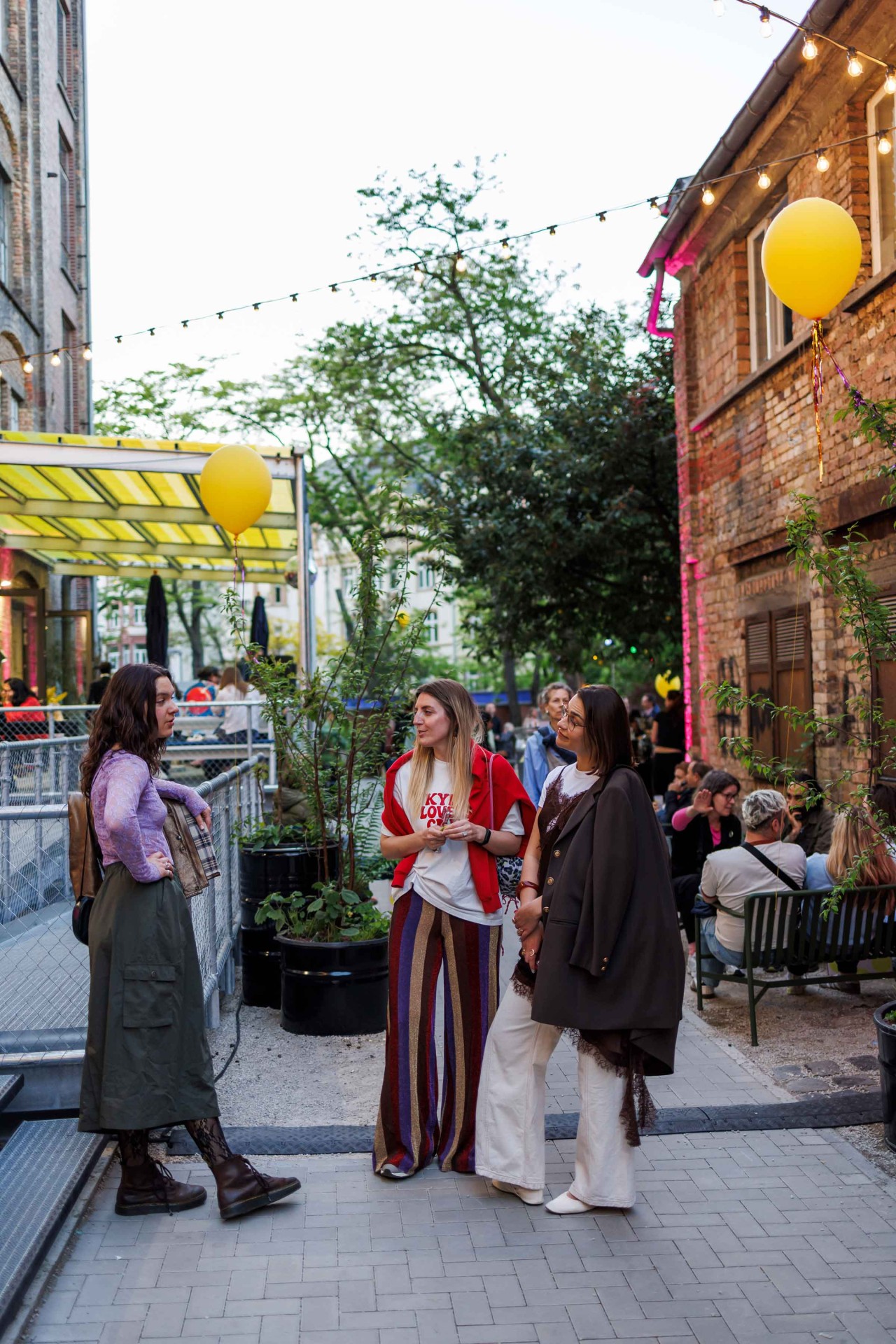 Three women outdoors in an urban setting. Colorful clothing, yellow balloons, and a relaxed atmosphere.