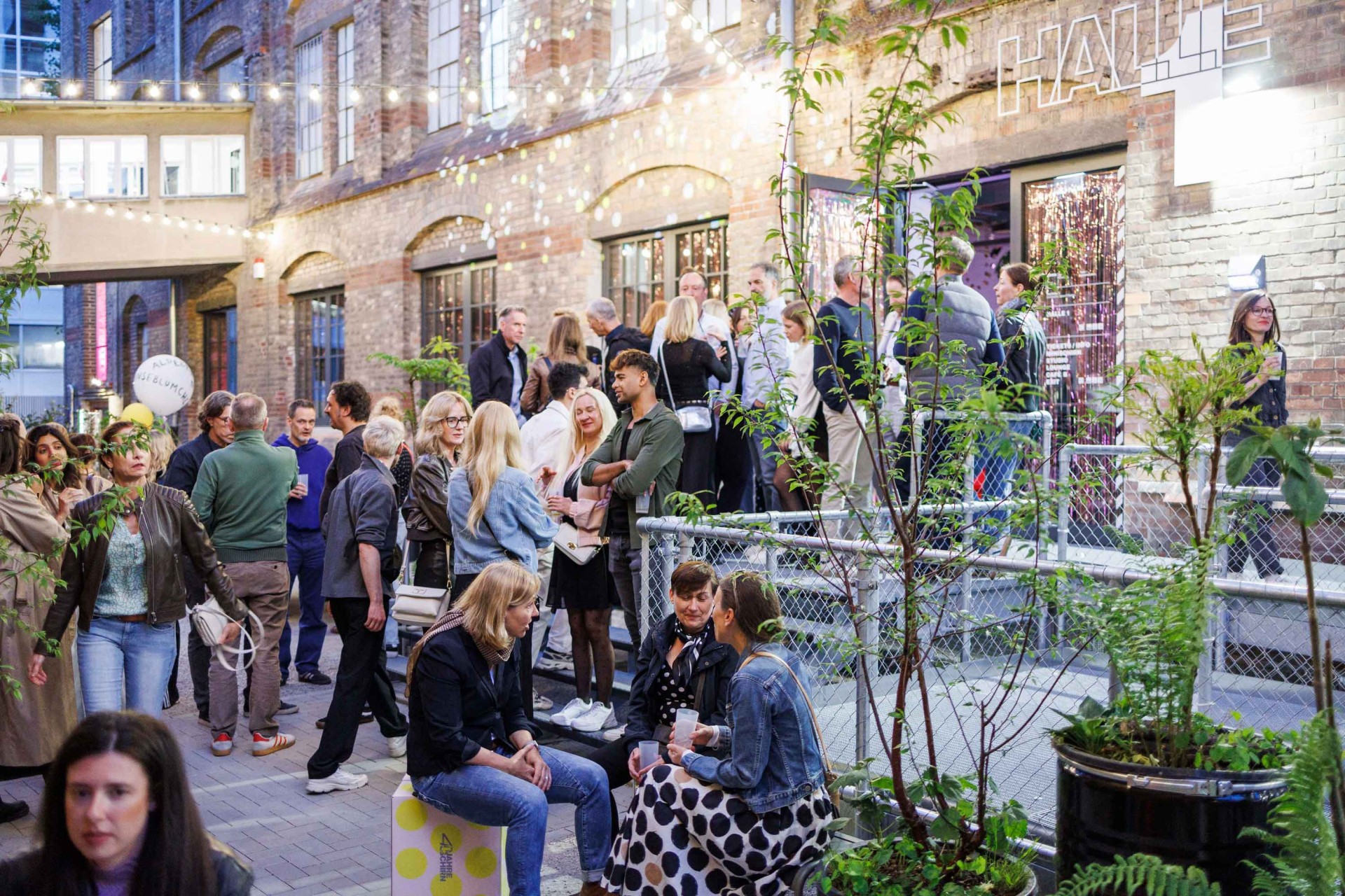 People gather in a lit courtyard, surrounded by plants and historic buildings.