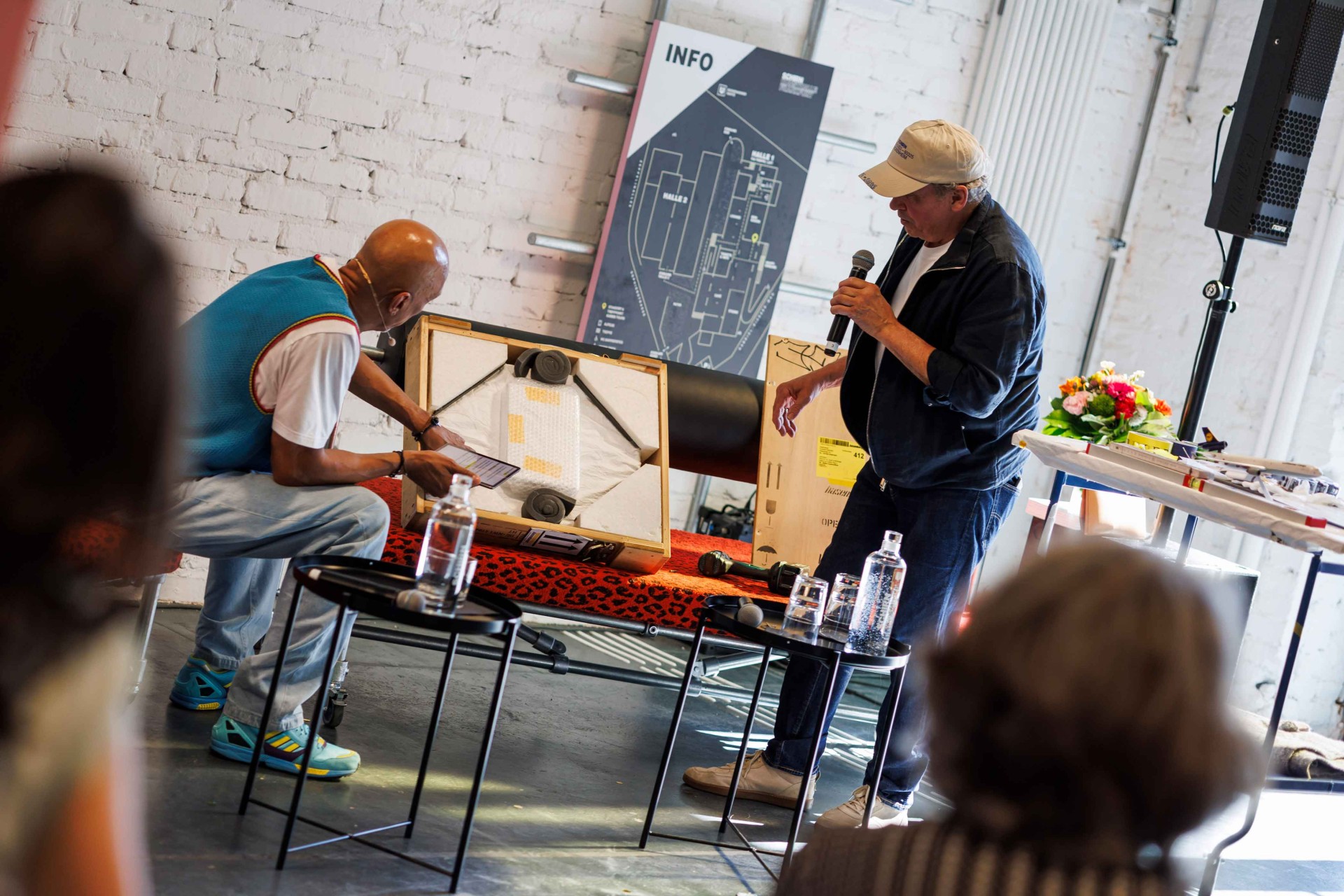 Two people discuss at a table with visible packaging and water bottles. An information poster is in the background.