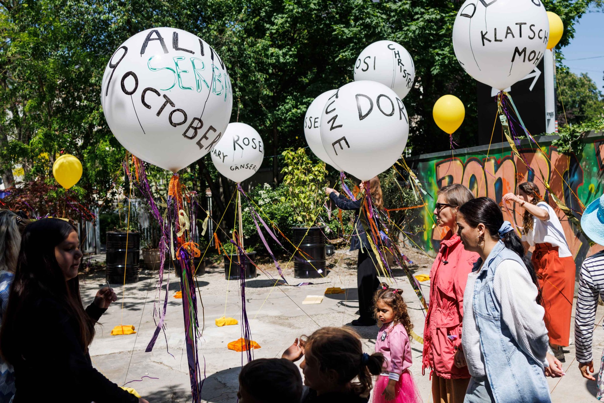 Group of people with balloons in a colorful, festive outdoor area under trees.