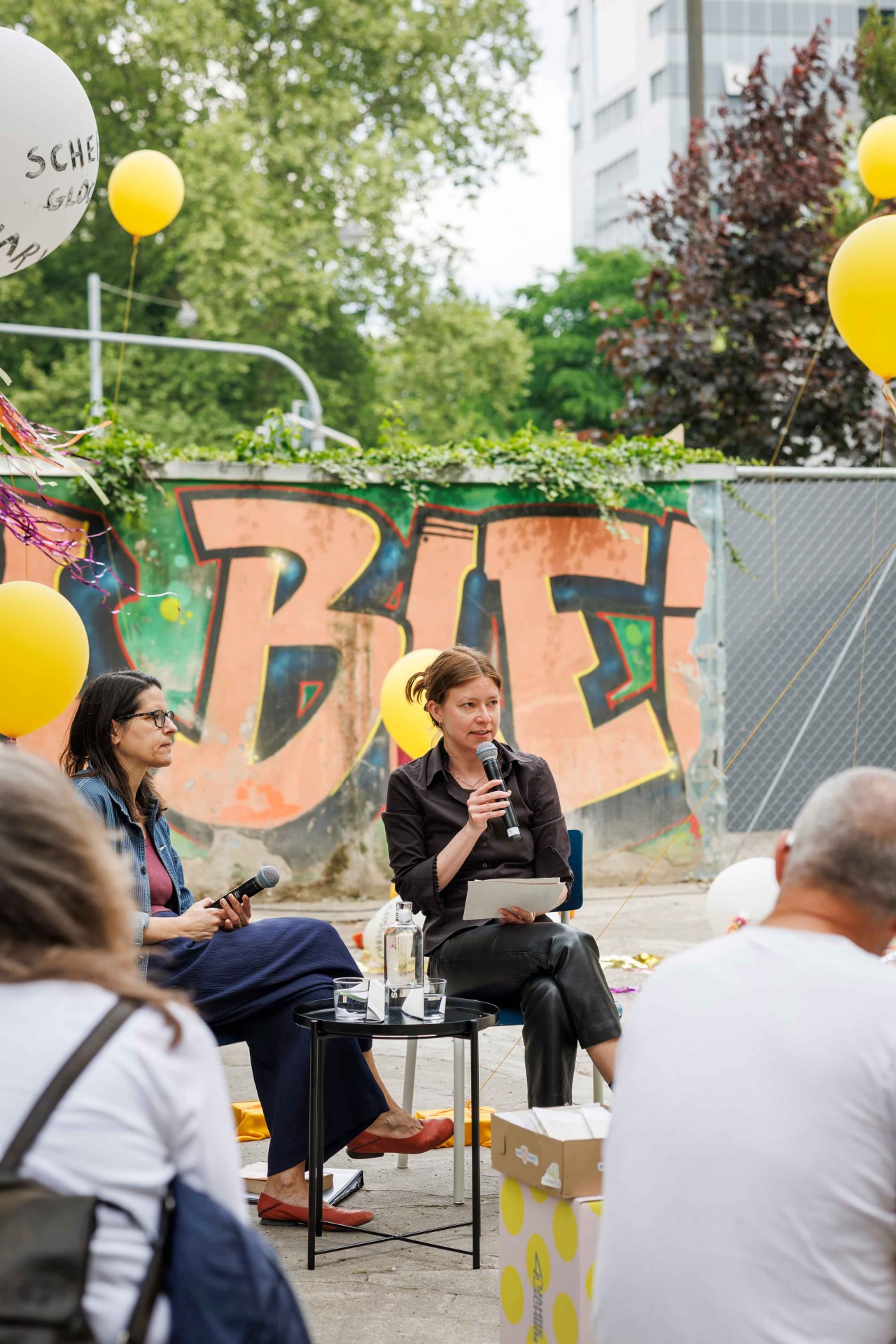 Two women are speaking at an event, surrounded by yellow balloons and a colorful graffiti wall in the background.