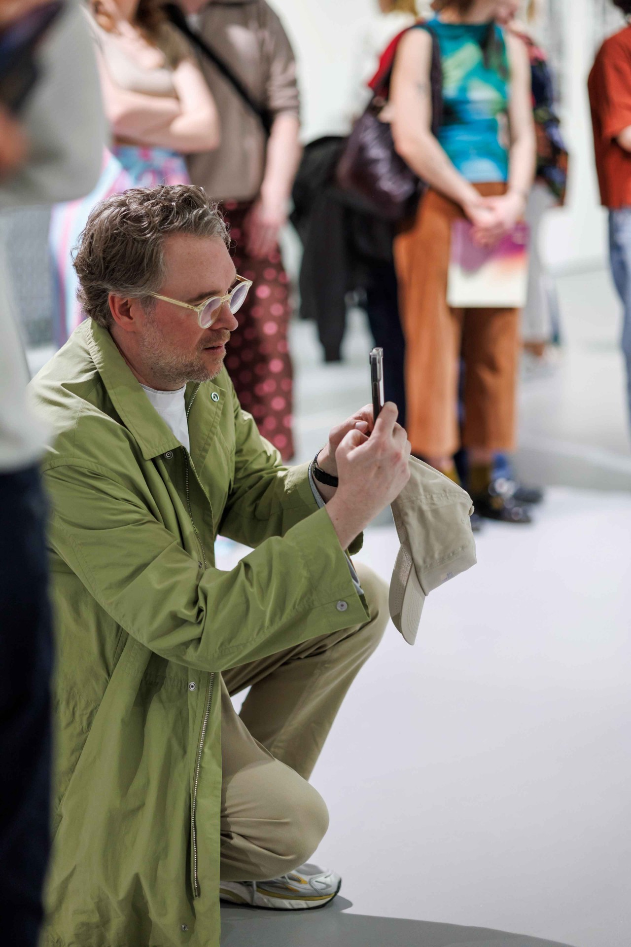 A man in a green coat kneels and takes a photo while many people stand in the background.