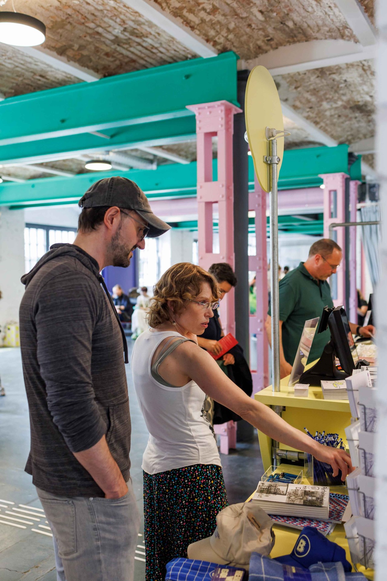 Two people are looking at various products at a sales booth, with a colorful space and other visitors in the background.