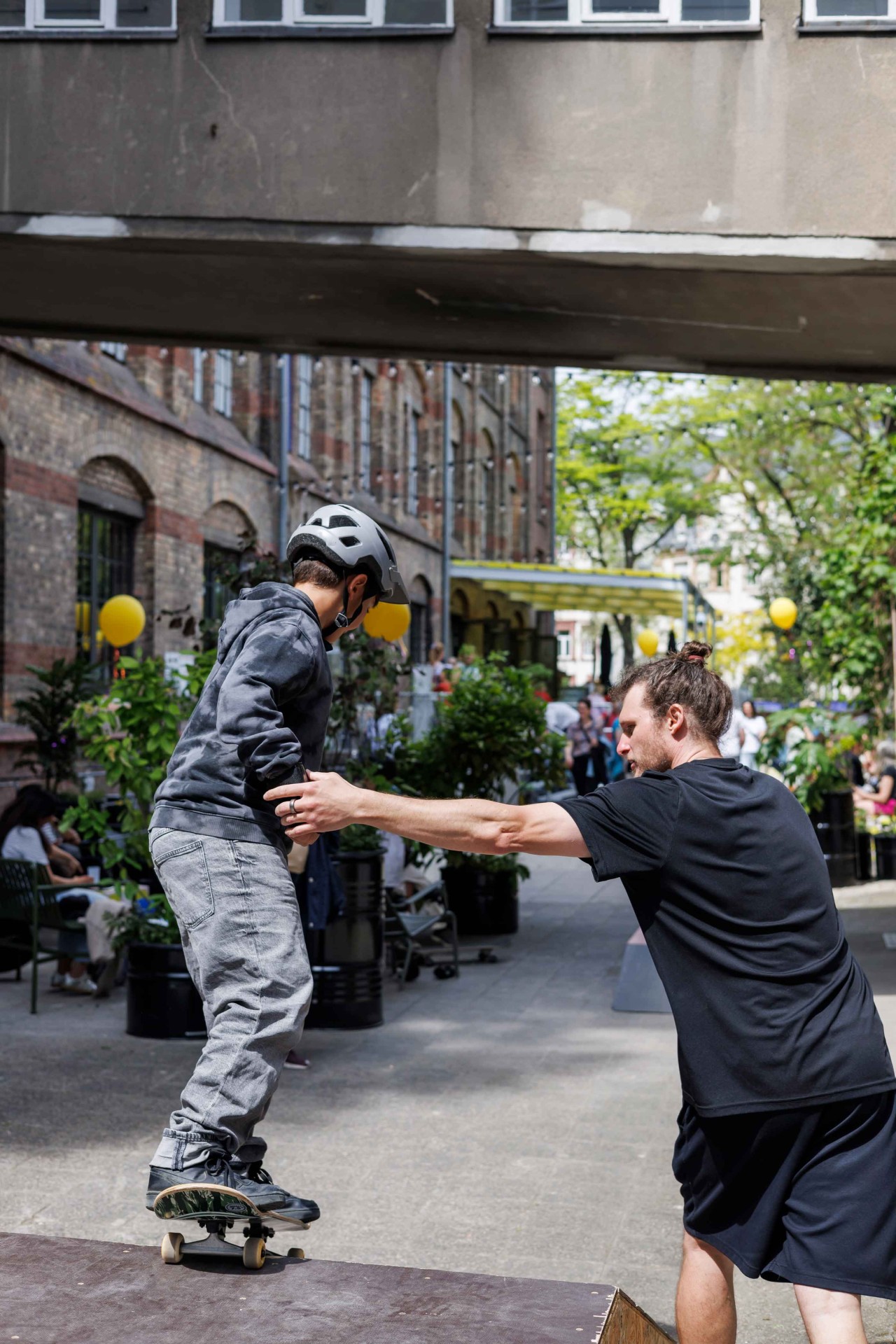 A boy in a helmet rides a skateboard while an adult supports him. In the background, there are people and plants visible.