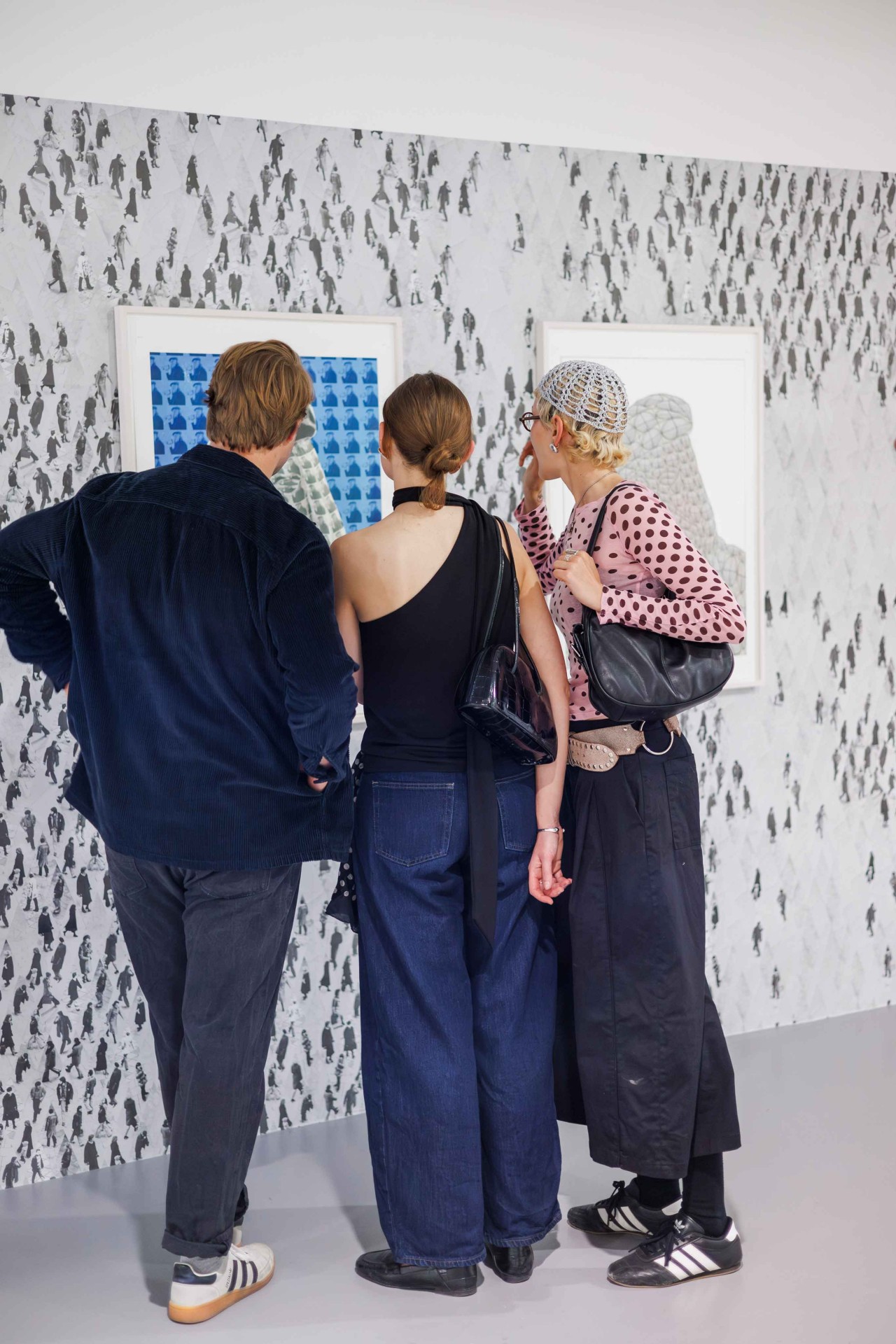 Three people are admiring artworks in front of a patterned wall in a modern exhibition space.