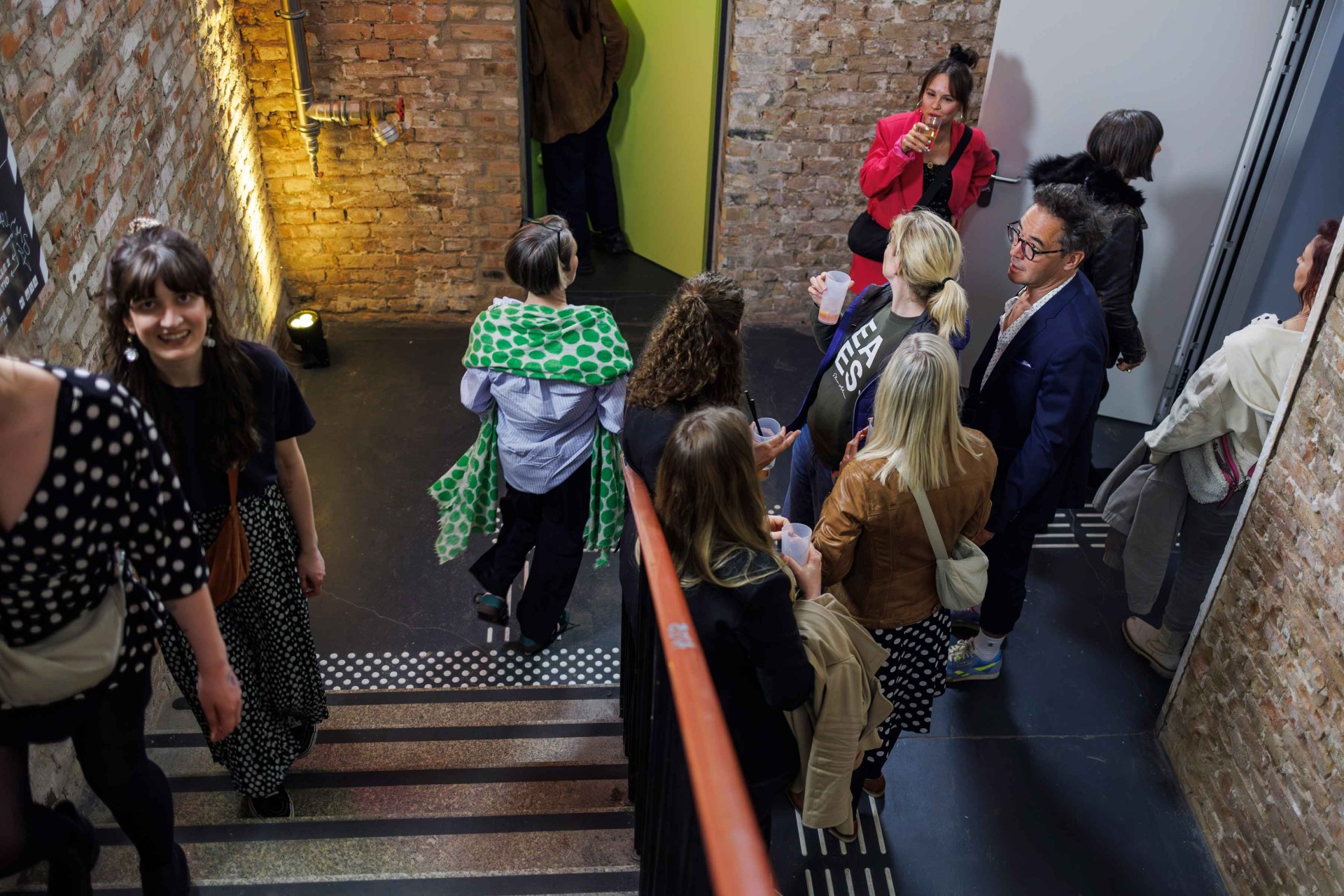 Guests are chatting in a modern room with brick walls and warm lighting, while some are going up the stairs.