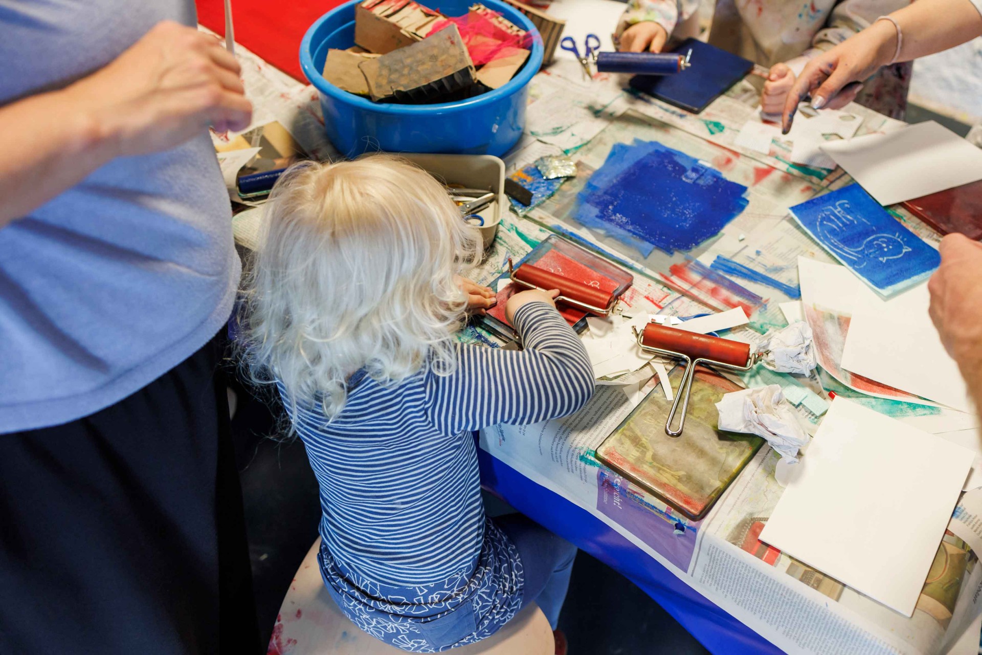 Child with long, blonde curls sits at a table full of art supplies, enthusiastically crafting.