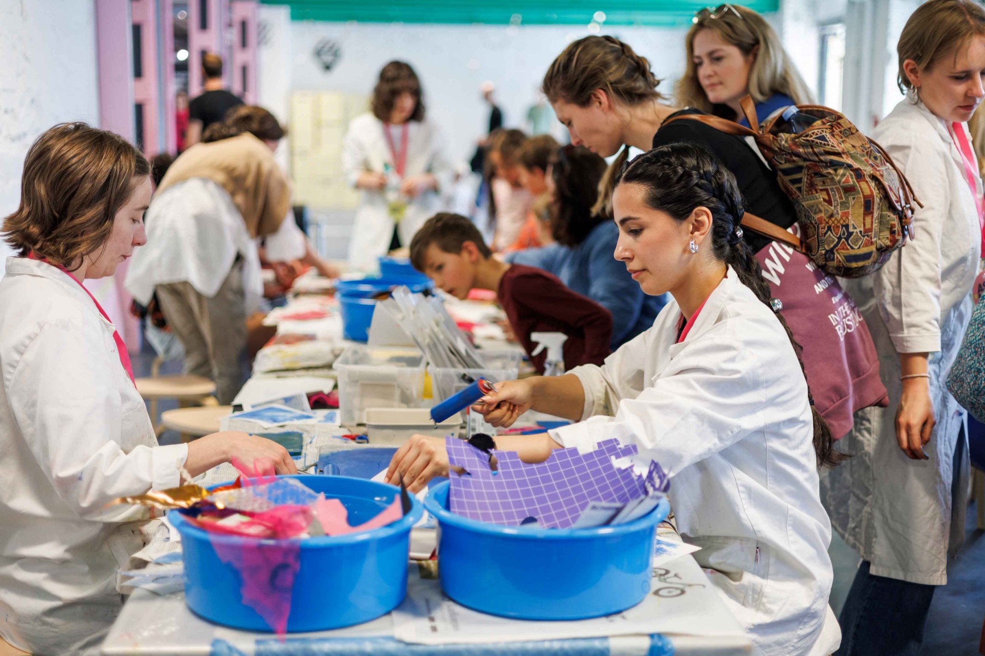 Various people work at tables, creatively using colorful materials and tools in a bright room.