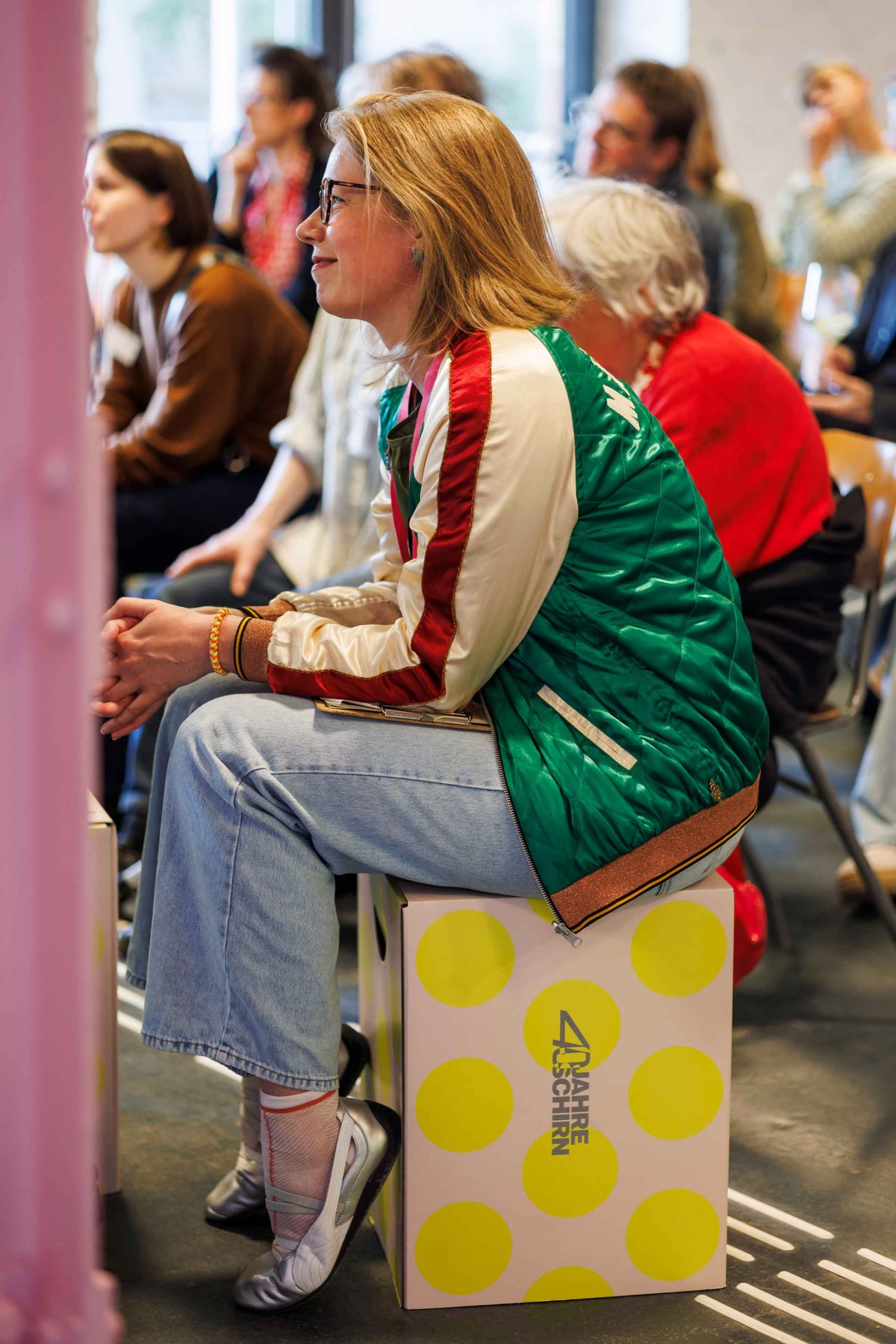 A woman with glasses sits on a box, wearing a green jacket and looking thoughtfully around.