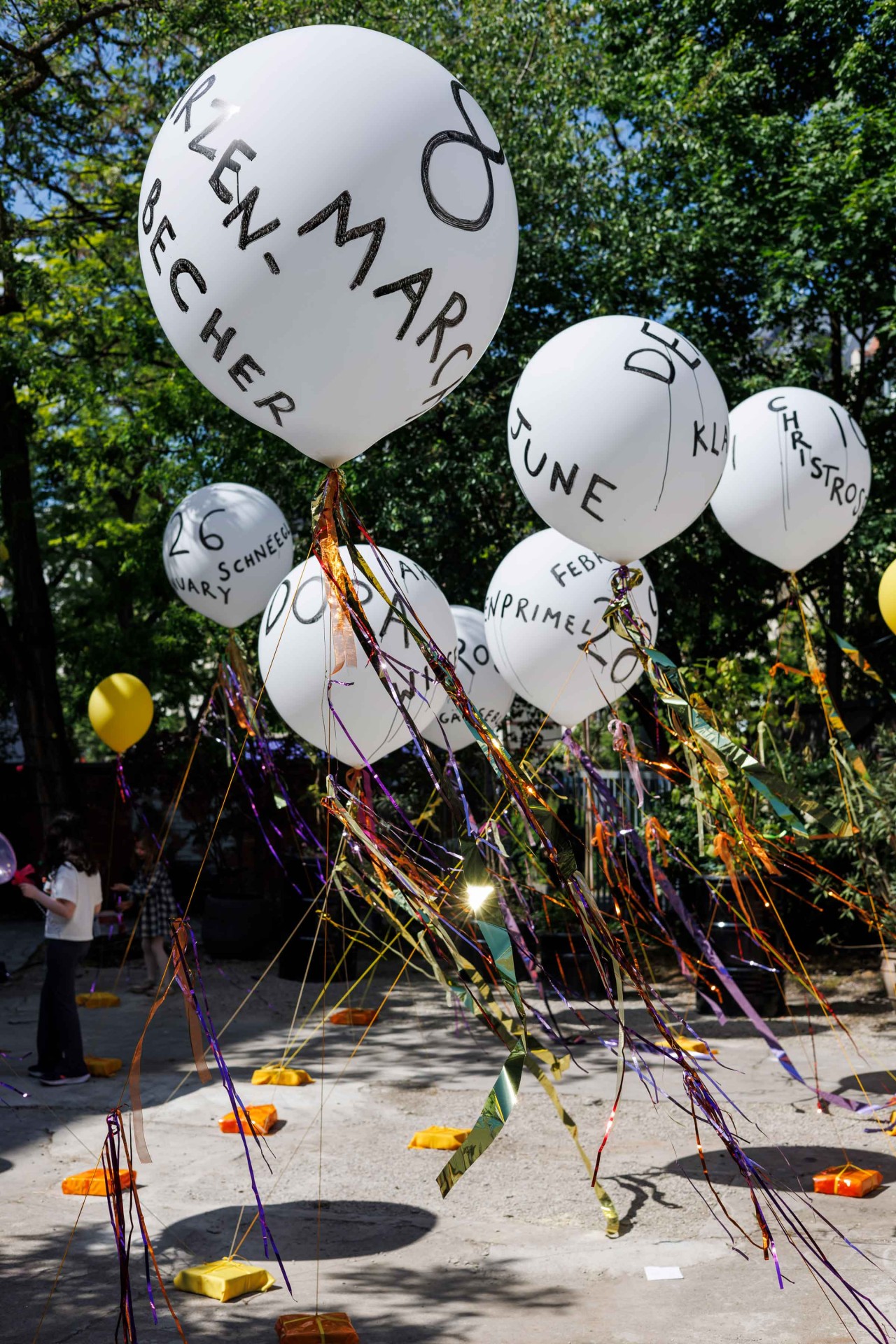 Colorful balloons with various names and dates, surrounded by vibrant ribbons in a sunny park.