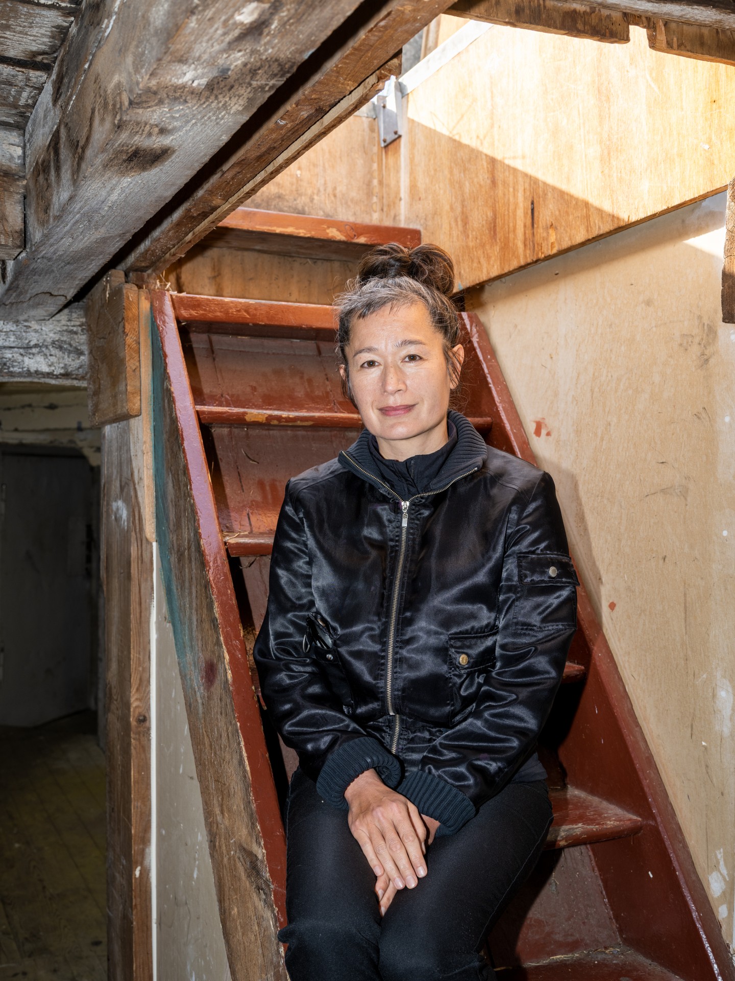 Eine Frau in schwarzer Jacke sitzt lächelnd auf einer Treppe in einem hellen Raum mit Holzverkleidung.