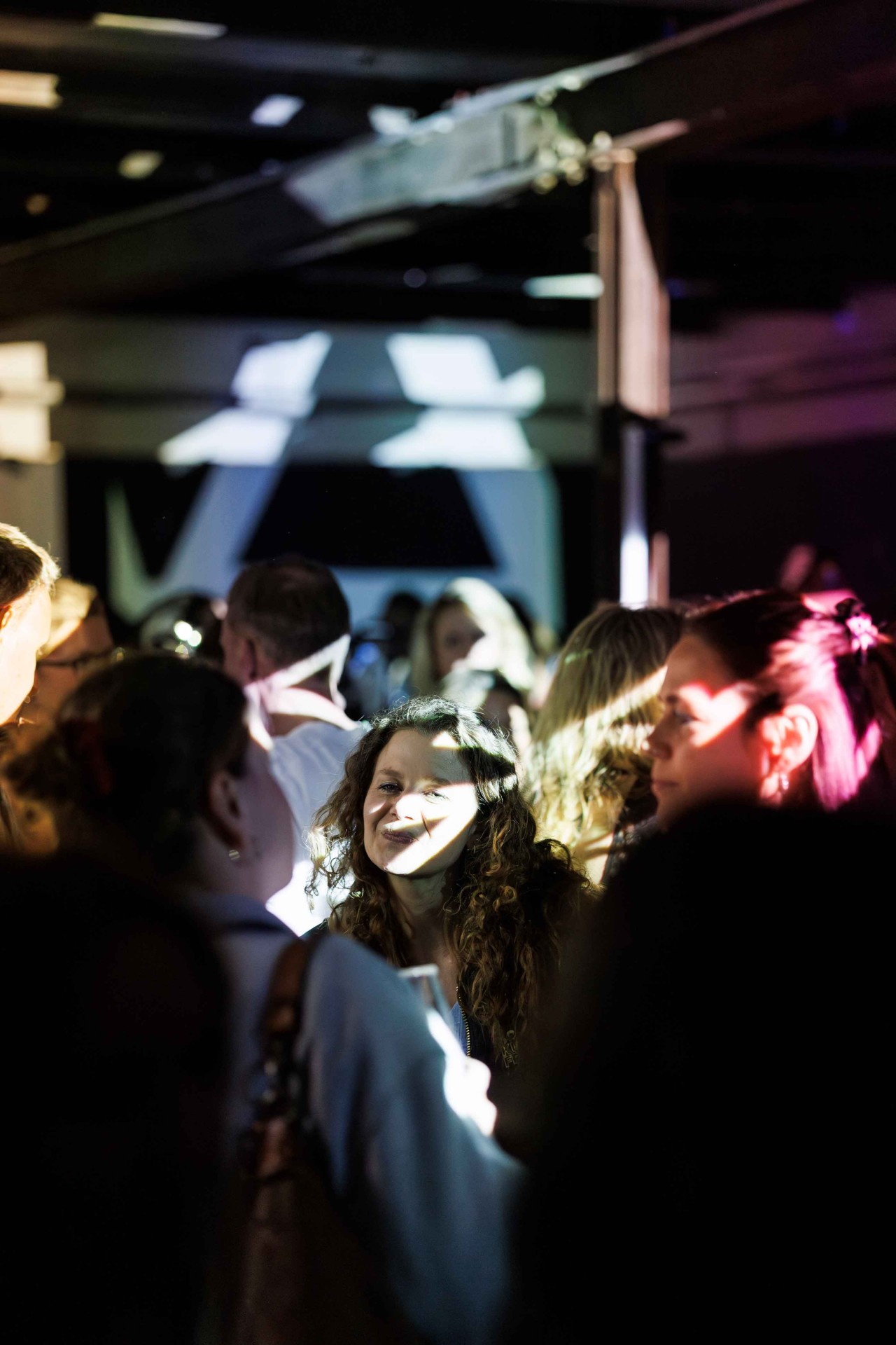 A crowd at an event, with a smiling person in focus and colorful lights in the background.