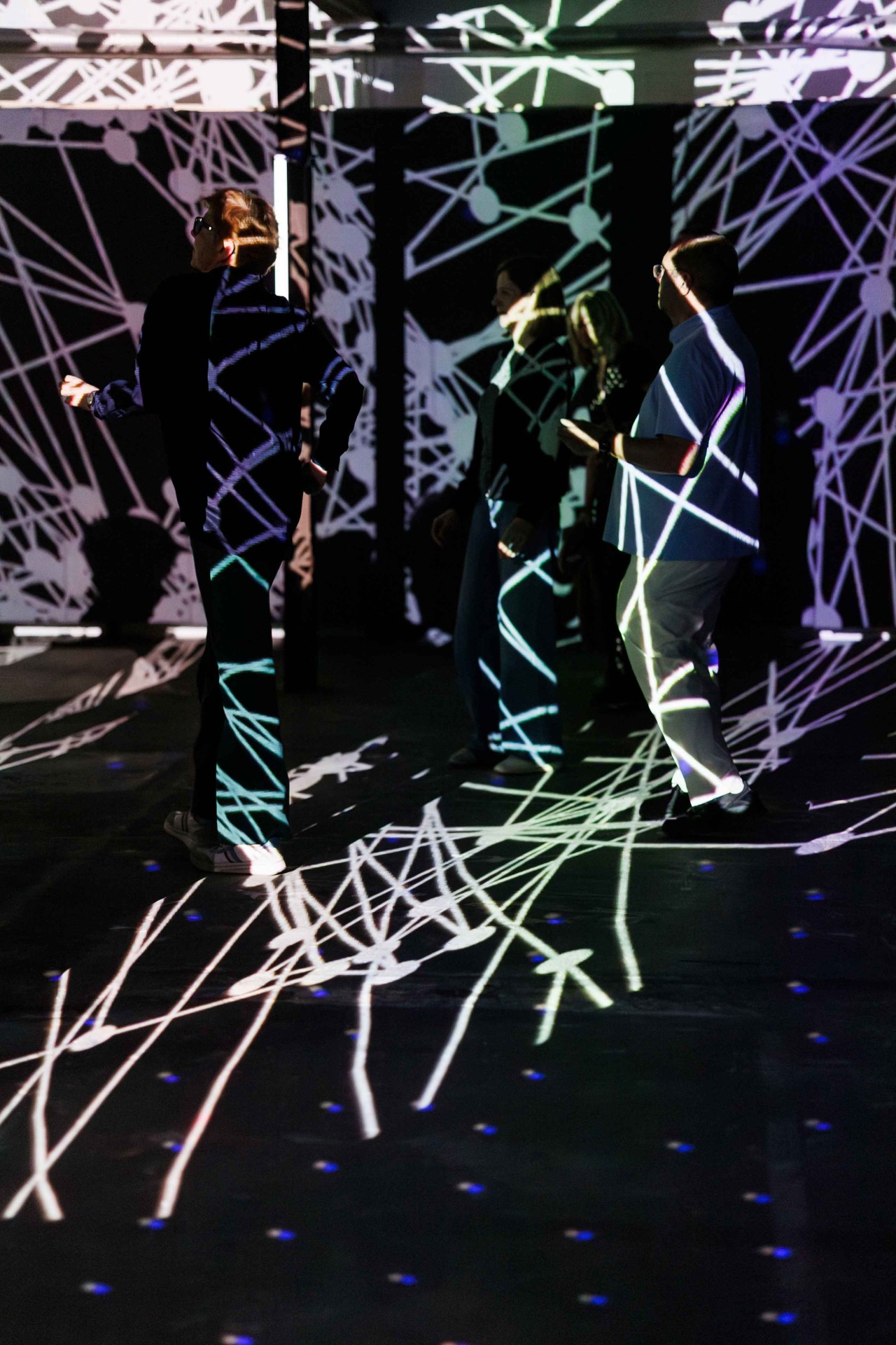 People move in a room with fascinating light projections and geometric patterns on the floor.