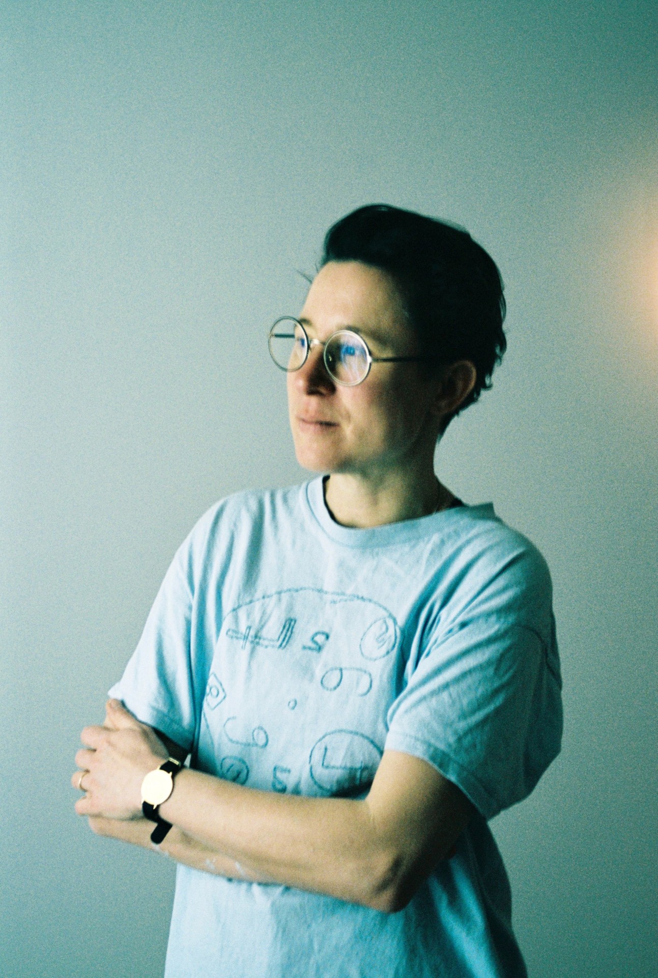 Person mit Brille in blauem T-Shirt, stehend mit verschränkten Armen, vor hellem Hintergrund.